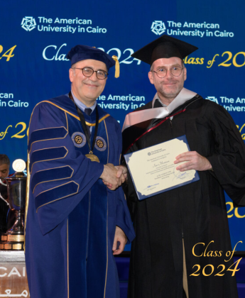 A professor in a cap and gown stands holding a certificate shaking hands with the President of AUC