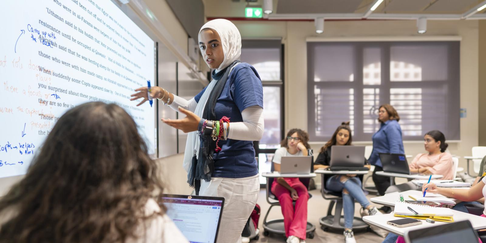 veiled student writing information infront of class on white board