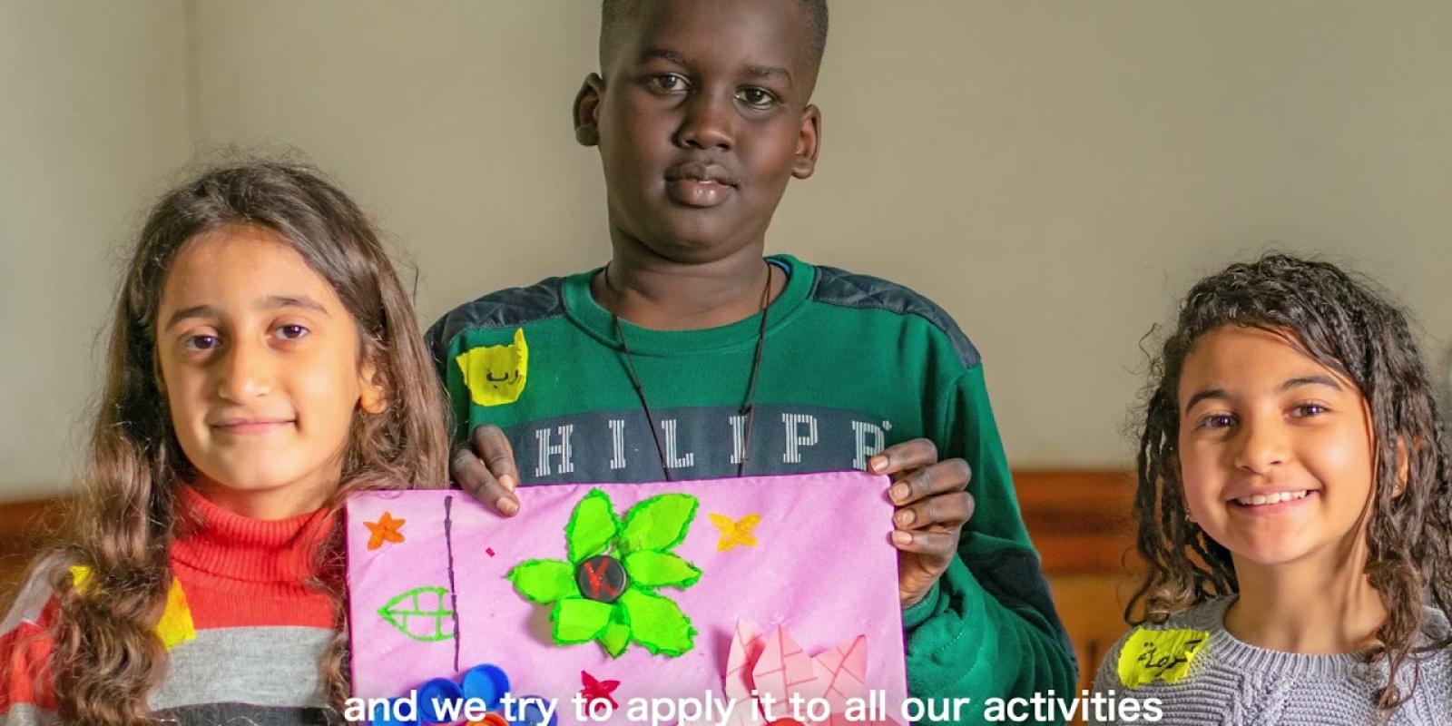 A boy and two girls are smiling. The boy is holding a colorful picture of green, pink and blue flowers