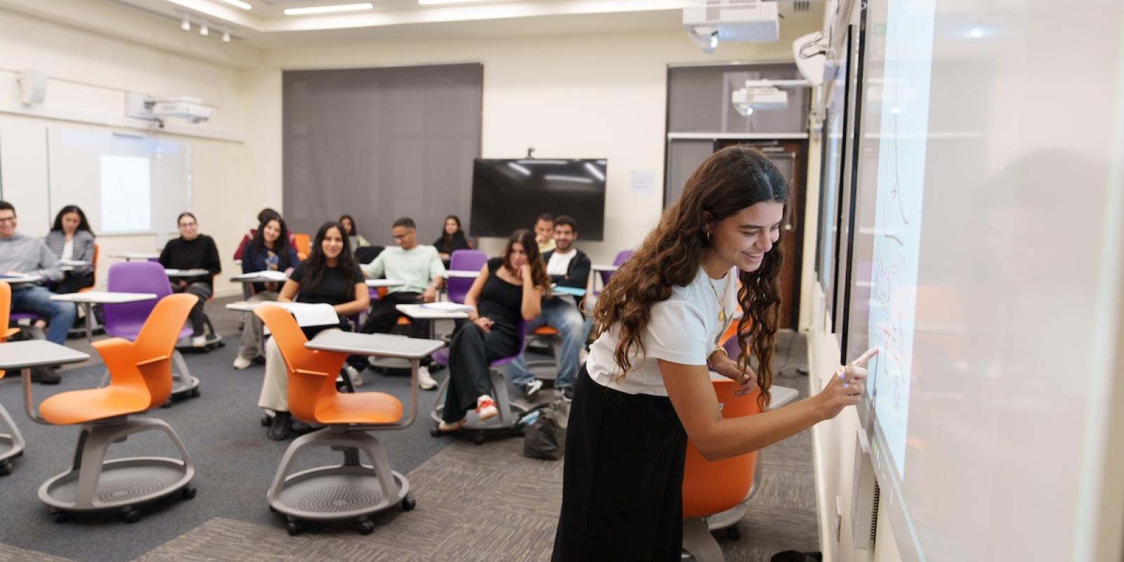 A student smiles while writing on a digital classroom board as classmates sit behind her in a interactive style classroom with colorful chairs.