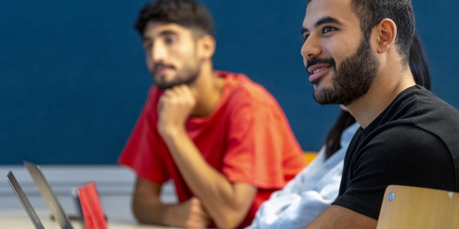 Two students sit at a table with laptops, listening attentively during a classroom discussion while one smiles and looks toward the speaker.