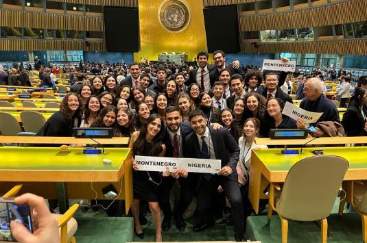 The CIMUN team stands wearing suits and professional dresses in an auditorium at the conference. Some students are holding signs saying "Montenegro" and "Nigeria"