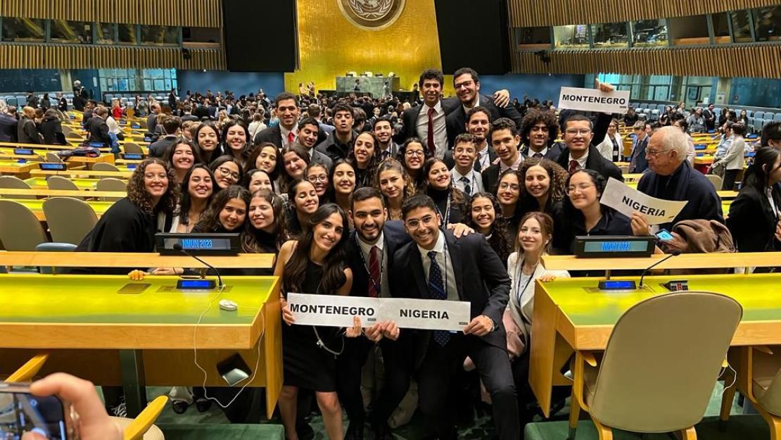 The CIMUN team stands wearing suits and professional dresses in an auditorium at the conference. Some students are holding signs saying "Montenegro" and "Nigeria"