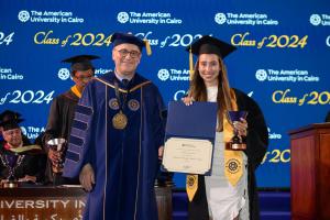 A woman in a cap and gown stands next to the President of AUC holding a certificate and a golden trophy