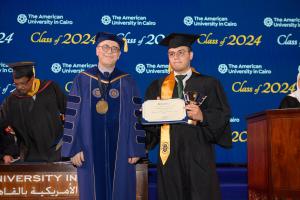 A man in a cap and gown stands next to the President of AUC holding a certificate and a golden trophy